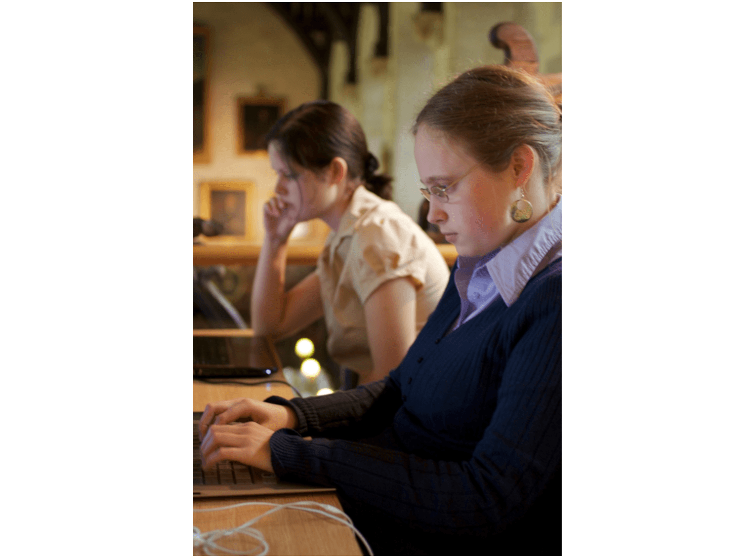 L'image montre deux femmes qui bossent sur des ordinateurs dans un endroit qui ressemble à un bureau ou une bibliothèque. La lumière est tamisée et l'ambiance est calme.