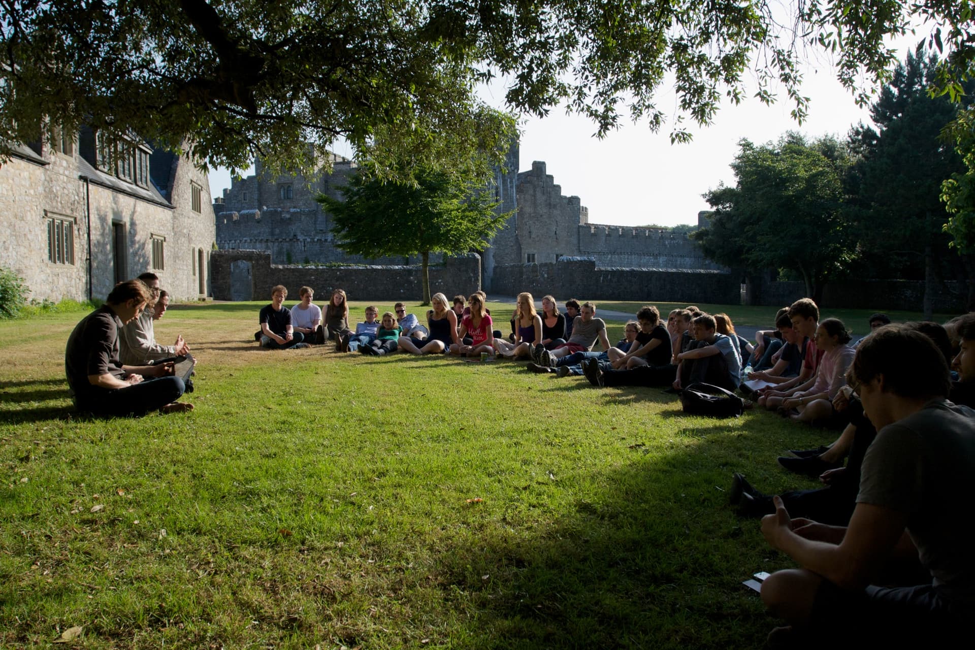 Un groupe de personnes sont assises en cercle sur une pelouse verte, en train d'écouter un orateur dans un décor qui ressemble à un campus historique ou à un parc avec de vieux bâtiments en pierre en arrière-plan.