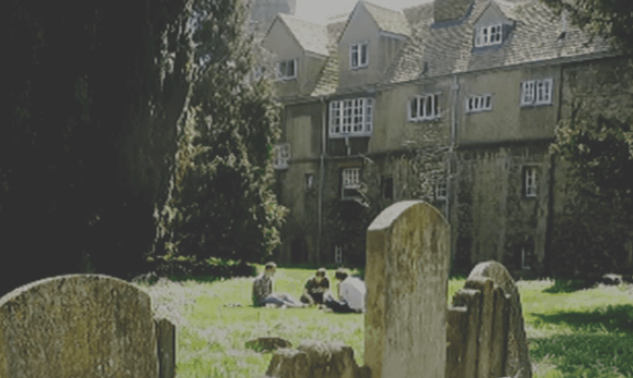 La photo montre un groupe de personnes assises sur la pelouse devant un vieux bâtiment, avec des pierres tombales au premier plan.
