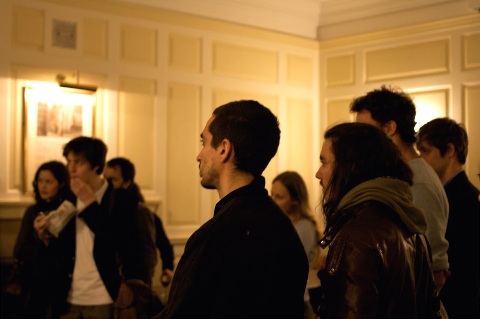 L'image montre un groupe de personnes dans un espace intérieur, peut-être lors d'un événement social ou d'une réunion, avec un jeune homme de profil au premier plan regardant vers la droite.