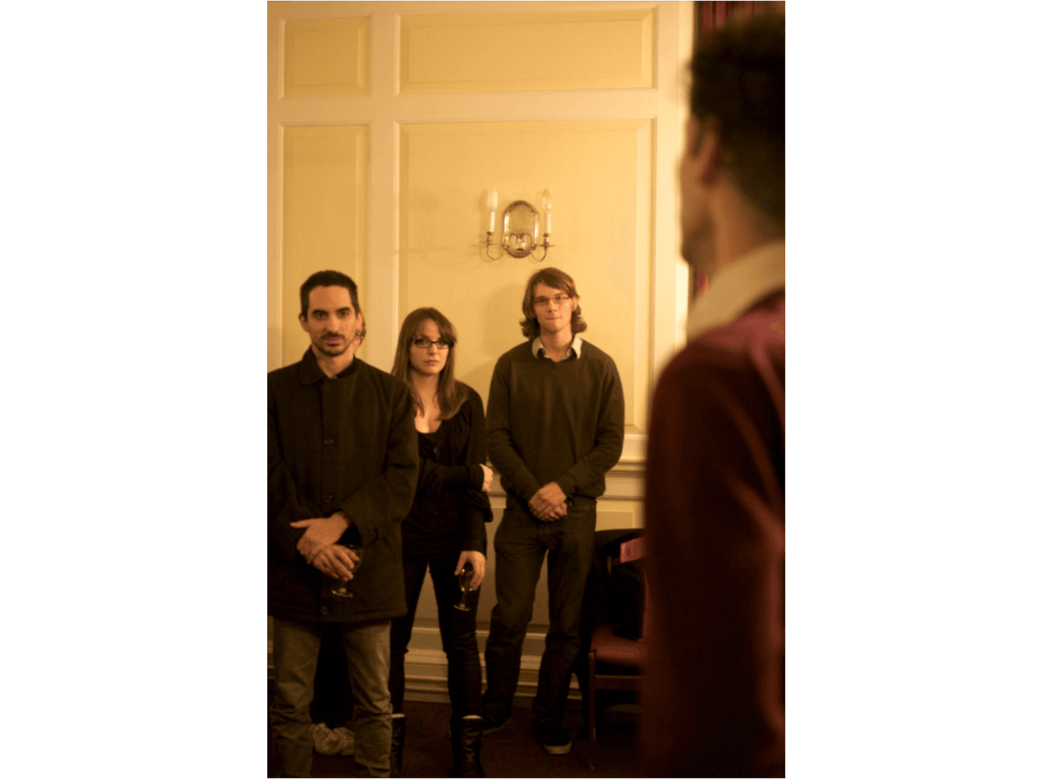 L'image montre trois personnes debout dans une pièce décorée de façon classique, regardant vers quelque chose ou quelqu'un hors champ, dans une ambiance détendue.