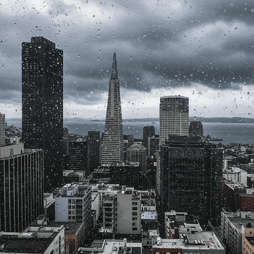 Une vue de la ligne d'horizon de San Francisco à travers une fenêtre couverte de gouttes de pluie. La ville semble grise sous un ciel nuageux, avec la baie visible au-delà des bâtiments.