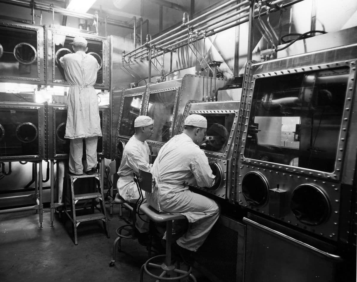 L'image montre trois personnes travaillant dans un laboratoire, toutes portant des vêtements de protection et manipulant du matériel à travers des compartiments étanches avec des gants intégrés.