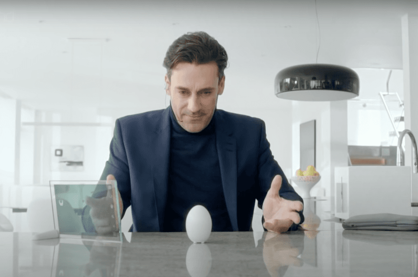 Un homme en costume se penche sur une surface polie dans une cuisine moderne, regardant avec intérêt un appareil en forme d'œuf positionné horizontalement. La scène suggère une atmosphère de curiosité ou de surprise.