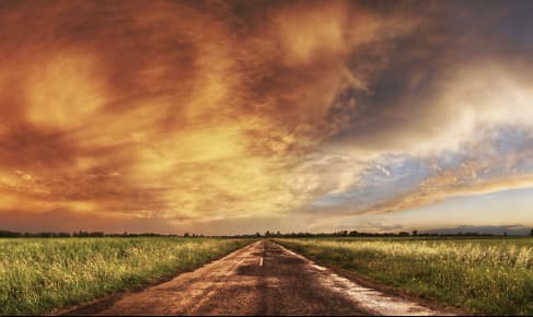 L'image montre une route droite traversant une plaine, bordée de champs verts sous un ciel qui paraît dramatique à gauche, avec des nuages reflétant des tons orange et jaune, et clair et lumineux à droite.
