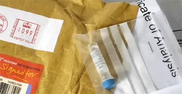 L'image montre un paquet ouvert avec une enveloppe en plastique transparent à l'intérieur, qui semble contenir un tube à essai ou un échantillonneur médical avec un couvercle bleu, suggérant la réception d'un composant ou d'un échantillon pour analyse.