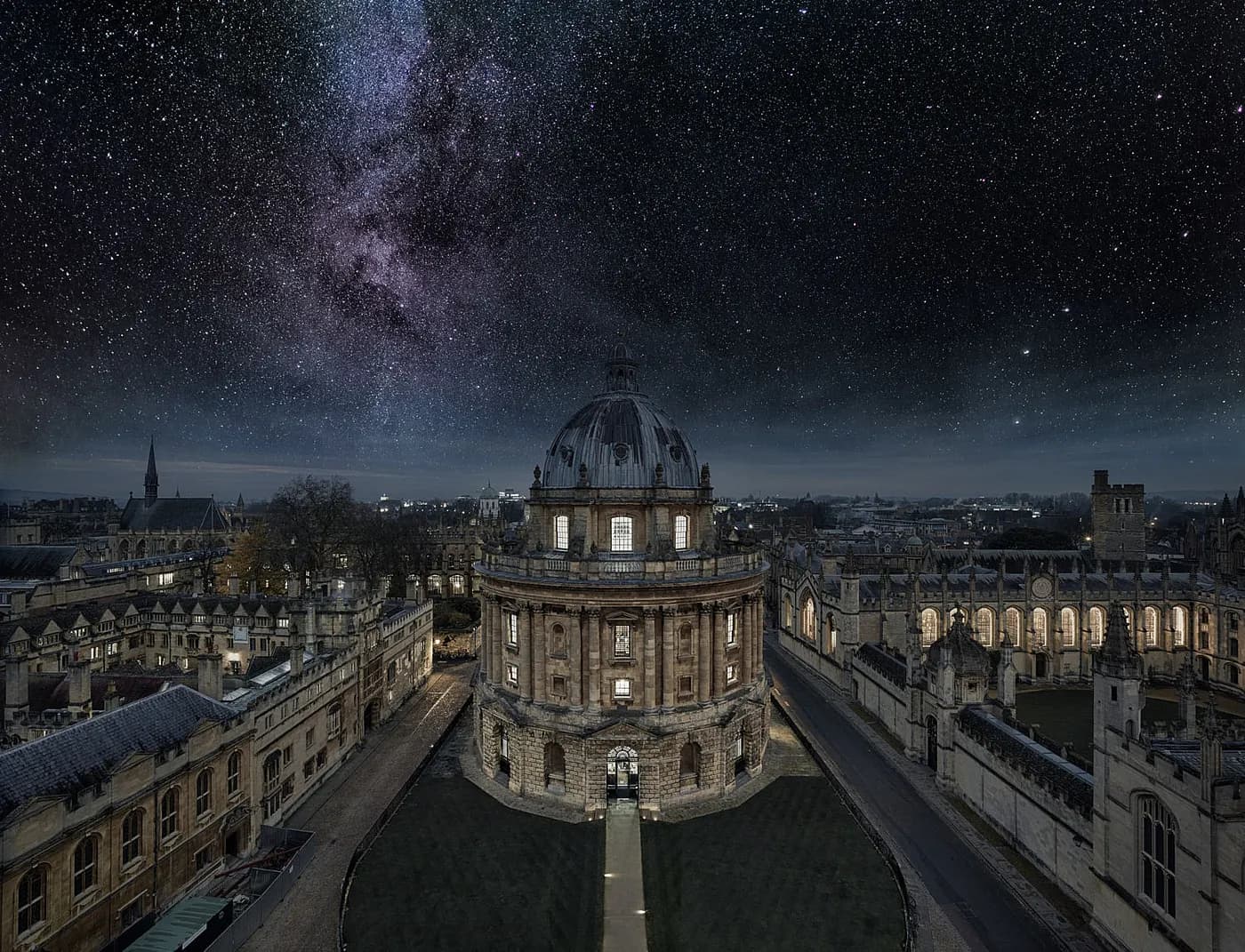 L'image montre une vue aérienne nocturne de la Radcliffe Camera, qui fait partie de la Bodleian Library de l'Université d'Oxford, avec un ciel étoilé et la Voie lactée visible en arrière-plan.